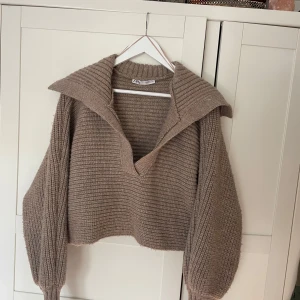 Beige stickad tröja med krage från Zara - Säljer en beige stickad tröja från Zara med bred krage och v-ringning. Tröjan har ribbade muddar och långa ärmar. Perfekt för lager-på-lager.