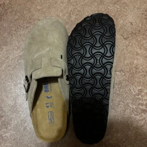 Mockasandaler från Birkenstock - Snygga sandaler från Birkenstock i beige mocka med rund tå och justerbart spänne i metall. Skorna har en platt sula i kork och svart gummi undertill för extra grepp. Perfekta för chill dagar och enkel stil.
