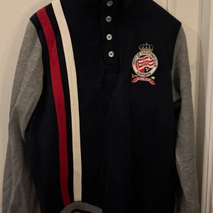 Marinblå pikétröja från Northern Basic - Snygg marinblå pikétröja från Northern Basic med grå ärmar, broderad logga på bröstet och coola röda och vita ränder framtill. Tröjan har knappar i halsen och sportig känsla, perfekt för dig som gillar preppy stil.