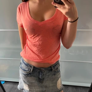 Korallrosa t-shirt från Hollister - Snygg korallrosa t-shirt från Hollister med v-ringning och korta ärmar. Liten vit logga broderad på bröstet. T-shirten är i mjuk bomull och har en avslappnad passform som är perfekt till jeans eller kjol.