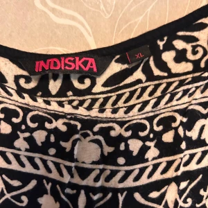 Svartvit mönstrad klänning Indiska XL - Svart klänning från Indiska i storlek XL med vitt, detaljerat blommönster och breda bårder. Ärmlös modell i mjuk viskos som faller snyggt. Perfekt för dig som gillar boho-vibbar och vill ha något unikt i garderoben.