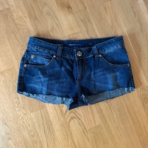 Miss Sixty Collection Jeansshorts - Snygga mörkblå jeansshorts från Miss Sixty med låg midja och rå, fransig kant nertill. Klassisk femficksmodell med bälteshällor och slitna detaljer för en cool look. Perfekta för varma dagar och festivaler.