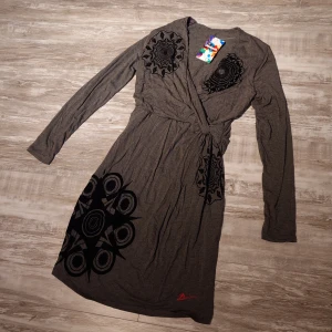 Grå omlottklänning från Desigual (Oanvänd) - Snygg grå omlottklänning från Desigual med långa ärmar och coola svarta grafiska mönster. Klänningen har knyt i midjan och v-ringad hals. Perfekt för dig som gillar unika detaljer och bekväm stil.