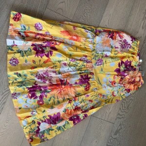 Gul blommig långkjol från H&M - Färgglad långkjol från H&M med blommigt mönster i lila, orange, grönt och vitt på en gul botten. Kjolen har resår i midjan och är tillverkad i mjuk bomull. 