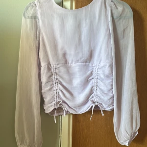 Lila blus från Lindex i storlek 164 - Superfin ljuslila blus med transparenta puffärmar och snygg snörningsdetalj framtill. Blusen har smockad rygg. Blusen är tajt i midjan och vid ryggen och lösare upptill.