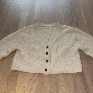 Beige ribbad kofta med knappar - Supermysig beige kofta med ribbad struktur och fyra mörka knappar framtill. Koftan har v-ringning och långa ärmar. Den är croppad och har en enkel, stilren look som passar till det mesta.