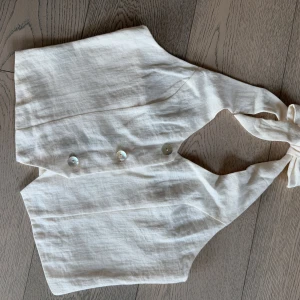 Beige topp från H&M i linne - Säljer en stilren beige topp från H&M i linne med omlottknut i nacken och två knappar framtill.