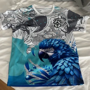 Nike CBF fotbollströja med blå papegoja - Unik fotbollströja från Nike med CBF-emblem, tillverkad i Dri-FIT-material. Tröjan har korta ärmar och ett färgstarkt tryck med en blå papegoja, blommor och grafiska detaljer i vitt, svart, grått och blått. Perfekt för dig som vill sticka ut på planen eller läktaren.