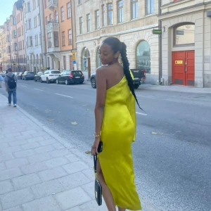Gul långklänning med oneshoulder - Snygg gul långklänning i satin med oneshoulder-design och en lång, vid ärm. Klänningen har slits på sidan och en elegant, figurnära passform. Perfekt för dig som vill sticka ut med en färgstark och trendig look.