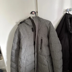 Grå pufferjacka från Take Off  - Säljer en grå pufferjacka från Take Off i storlek L. Jackan har huva, dragkedja framtill och en bröstficka med dragkedja. Insidan är fodrad med mjuk fleece för extra värme. Perfekt för kalla dagar och har en clean, stilren look.