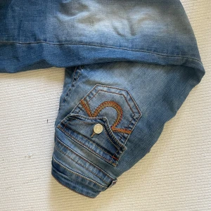 True Religion bootcut jeans blå - Säljer ett par blå bootcut jeans från True Religion med ikoniska bakfickor och kontrastsömmar i orange. Jeansen har låg midja, klassisk femficksdesign och snygg slitning framtill. Perfekta för dig som gillar en avslappnad men ändå trendig look.