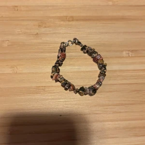 Armband med färgade stenar - Unikt armband med oregelbundna stenar i nyanser av rosa, brun, beige och svart. Stenarna är trädda på en tråd och armbandet har ett metallspänne. Perfekt accessoar för dig som gillar boho-stil och naturliga material.