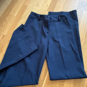 Mörkblå kostymbyxor med raka ben - Snygga mörkblå kostymbyxor med klassisk rak passform och hällor för bälte. Perfekta för en clean och stilren look. Byxorna har diskreta fickor och är tillverkade i ett mjukt tyg som känns bekvämt hela dagen.