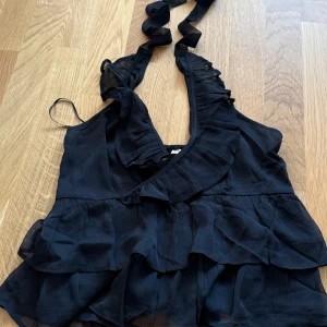 Svart volangig omlottopp - Supersöt svart omlottopp med volanger och djup v-ringning. Toppen har knytband i nacken och är ärmlös, perfekt för en trendig look. Tunt, lätt tyg som ger en luftig känsla och snyggt fall.