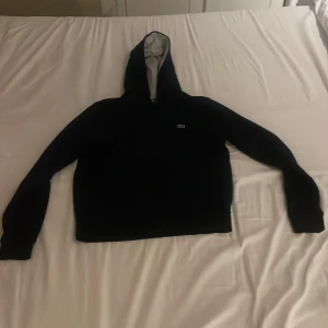 Mörkblå hoodie från Lacoste Sport - Mörkblå hoodie från Lacoste Sport med dragkedja och klassisk krokodillogga på bröstet. Tröjan har huva med grått innerfoder och långa ärmar. Perfekt för dig som gillar sportig och clean stil.