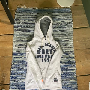 Vit hoodie från Superdry med tryck - Vit hoodie från Superdry med mörkblå texttryck framtill och klassisk känguruficka. Tröjan har huva med snörning och mjukt fleecefoder på insidan. Perfekt för dig som gillar sportig och avslappnad stil.   Hoodien har 2 fläckar.