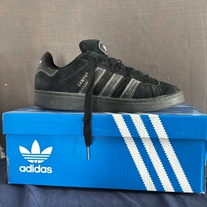 Adidas Campus 00s Svarta Sneakers - Adidas Campus 00s sneakers i svart mocka med klassiska vita ränder och logga på hälen. Skorna har rund tå, platt sula och snörning. Perfekt för dig som gillar retrovibbar och streetstyle. Säljs med originalkartong.