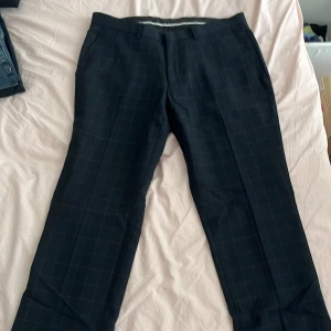 Mörkblå rutiga kostymbyxor från bläck - Snygga mörkblå kostymbyxor med diskret rutmönster i ljusblått. Klassisk rak passform med hällor för bälte och fickor både fram och bak. Perfekta för en clean och stilren look. Materialet känns mjukt och följsamt.
