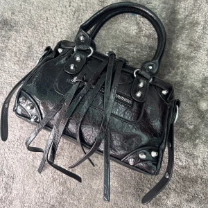 Svart handväska med nitar och dragkedja - Cool svart handväska i skinn med silverfärgade nitar, dragkedja framtill och långa dekorativa remmar. Väskan har två handtag och detaljer som ger en edgy vibe. Perfekt för dig som gillar statement-accessoarer. Aldrig använd, finns axelband också 