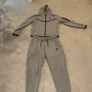 Grå mjukisbyxor från Nike med snörning - Gråa mjukisbyxor från Nike med svart logga på benet och snörning i midjan. Byxorna har en relaxed passform med muddar vid bensluten och är tillverkade i mjukt bomullsmaterial. Perfekta för chill eller träning.