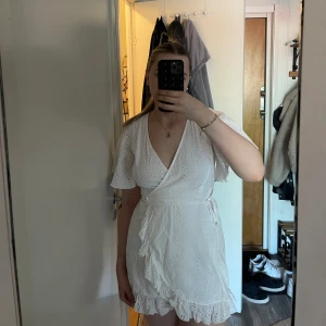 Vit omlottklänning från NA-KD - Supersöt vit kort omlottklänning från NA-KD med broderade detaljer och volangkant. Klänningen har v-ringning, knyt i midjan och luftiga korta ärmar.