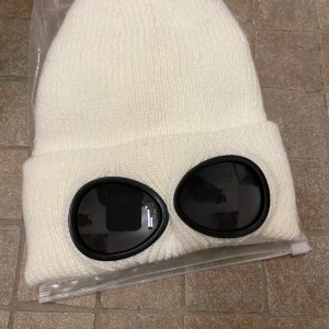 Vit ribbstickad mössa med goggles - Cool vit ribbstickad mössa med svarta goggles framtill. Mössan har ett mjukt och fluffigt foder på insidan som gör den extra varm och skön. Perfekt för dig som vill sticka ut med en unik accessoar i vinterkylan.