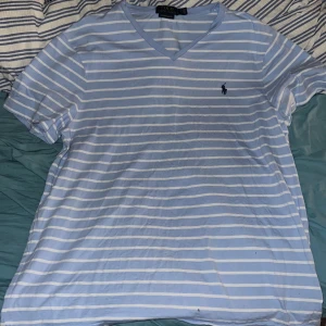 Blåvit randig t-shirt Polo Ralph Lauren - Snygg blå och vit randig t-shirt från Polo Ralph Lauren med broderad logga på bröstet. T-shirten har v-ringning, korta ärmar och är i en slim fit-modell. Tillverkad i 100% bomull för en skön och luftig känsla.