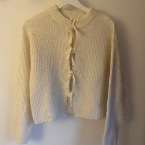 Krämvit kofta med rosettknäppning - Superfin krämvit kofta från ginatricot i ullmix med ribbstickad struktur. Koftan har långa ärmar och stängs med flera söta rosetter framtill. Perfekt för lager-på-lager och ger en mysig vibe till din outfit.