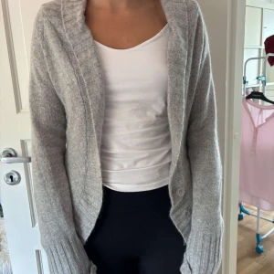 Grå kofta med bälte från ONLY - Mysig grå kofta från ONLY med bred ribbad kant, knappar framtill och matchande bälte i midjan. Koftan är stickad, har långa ärmar och en relaxed fit som passar perfekt till chill dagar. Enkel att styla över en t-shirt eller linne. Passar även M och S.