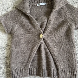 Beige stickad kofta från Zara - Kortärmad stickad kofta från Zara i beige nyans. Koftan har en stor krage, en dekorativ knapp framtill och ribbade kanter. Perfekt för lager-på-lager och chill vibes.
