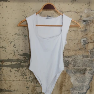 Vit ärmlös bodysuit från Zara - Snygg vit bodysuit från Zara med breda axelband och fyrkantig urringning. Perfekt att styla med jeans eller kjol för en clean och fräsch look. Mjukt och stretchigt material som sitter skönt mot kroppen.