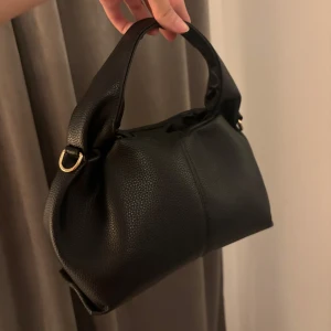 Svart handväska i skinn med gulddetaljer - Snygg svart handväska i skinn med mjuk struktur och guldfärgade metalldetaljer. Väskan har ett brett handtag och stilren design som passar till många outfits. Perfekt storlek för det viktigaste och enkel att bära i handen.