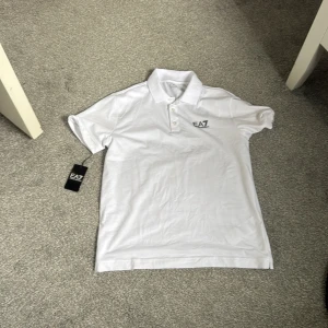 Vit pikétröja från Emporio Armani EA7 - Snygg vit pikétröja från Emporio Armani EA7 i storlek S. Klassisk design med krage, knappar framtill och diskret EA7-logga på bröstet. Tillverkad i mjuk bomull som känns skön mot huden. Perfekt för en clean och stilren look.