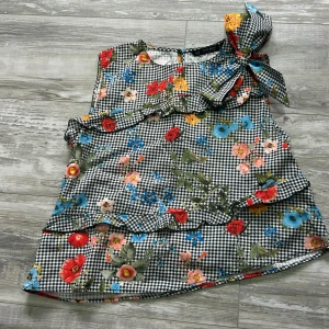 Blommig rutig blus från Zara Basic - Supersöt blus från Zara Basic med svartvitt rutigt mönster och färgglada blommor i rött, blått, gult och rosa. Toppen är ärmlös, har volangdetaljer och en stor rosett på axeln. Perfekt för dig som gillar unika och lekfulla plagg!