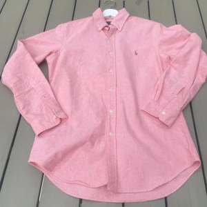Rosa skjorta från Ralph Lauren - Klassisk rosa skjorta från Ralph Lauren med button down-krage och broderad logga på bröstet. Skjortan har lång ärm, knäppning framtill och är tillverkad i bomull. Perfekt för en clean och stilren look. Hör av dig vid minsta fundering