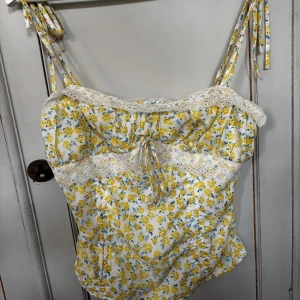 Blommig korsettopp med spets och snörning - Supersöt korsettopp med vita och gula blommor på vit botten, dekorerad med spetsdetaljer och snörning framtill. Axelbanden är smala och knyts upptill. Toppen är unik då jag sytt om den från en klänning. Passar storlek S