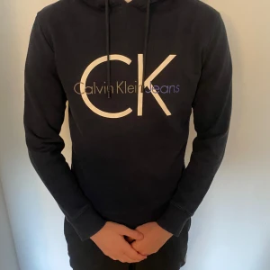 Svart hoodie från Calvin Klein Jeans - Snygg svart hoodie från Calvin Klein Jeans med stort CK-tryck på bröstet. Använd flitigt men är i bra skick! Säljer då den är blivit för liten 