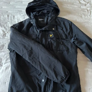 Svart vindjacka från Lyle & Scott - Svart vindjacka från Lyle & Scott med huva och klassisk gul logga på bröstet. Jackan har dragkedja och tryckknappar framtill, två bröstfickor och meshfoder på insidan. Perfekt för blåsiga dagar och enkel att matcha med allt.