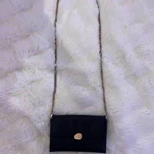 Svart väska med kedjerem från Glitter - Snygg svart kuvertväska med struktur i materialet och guldfärgad kedjerem. Väskan har ett stilrent guldspänne framtill som en knapp. Den har några defekter men inget som syns så mycket. Bara använd ett fåtal gånger. 