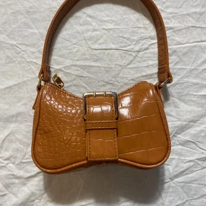 Brun axelväska med krokoprägling - Snygg brun axelväska i syntetmaterial med krokodilpräglad yta och coolt bältedetalj framtill. Väskan har en unik form, magnetlås och guldfärgade metalldetaljer. Perfekt för dig som vill sticka ut med accessoarerna.