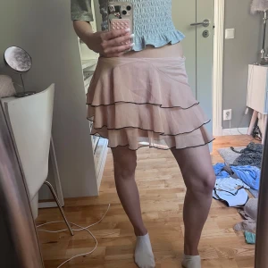 Beige volangkjol med svarta kanter - Supersöt beige kortkjol med flera lager volanger och svarta kanter. Kjolen har en lätt och flowy känsla, perfekt för dig som gillar en feminin och lekfull stil. Materialet känns tunt och luftigt, vilket gör den extra skön att bära varma dagar.