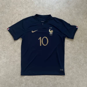 Frankrike hemmatröja/vm tröjan 2022/23 - Snygg marinblå Frankrike fotbollströja från Nike med guldiga detaljer, nummer 10 och Mbappé på ryggen. Tröjan har korta ärmar och är tillverkad i ett lätt och ventilerande material. Perfekt för dig som vill bära landslagets färger och hylla Mbappé.