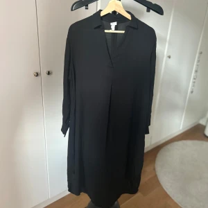 Svart långärmad skjortklänning - En svart, enkel och stilren skjortklänning från H&M med v-ringning och krage. Klänningen har lång ärm och en lös passform som ger en avslappnad vibe. Perfekt för dig som gillar minimalistisk stil.