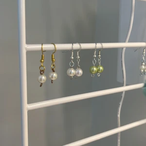 Pärlörhängen i olika färger - Säljer tre par handgjorda örhängen med pärlor. Ett par har guldfärgade krokar och vita pärlor, ett par har silverfärgade krokar och vita pärlor, och ett par har silverfärgade krokar med ljusgröna pärlor. Säljer 1 par för 35kr och alla 3 par för 80kr