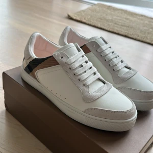 Burberry vita sneakers med rutigt mönster - Snygga vita sneakers från Burberry med klassiskt beige, svart och brunrutig panel på hälen. Skorna har ovandel i skinn och mockadetaljer vid snörningen och tån. Platt sula och rund tå ger en clean look. Perfekta för dig som vill ha en stilren men ändå lyxig sneaker.
