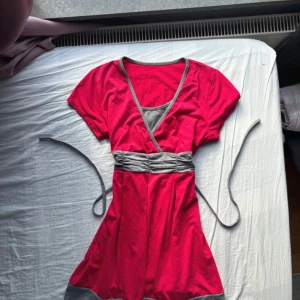 Röd omlotttopp med grå detaljer - Supersöt röd lång topp med grå omlottdetaljer och knytband i midjan. Toppen har korta ärmar, v-ringning och grå kant vid halsen samt nederkanten. Perfekt för dig som gillar färg och vill ha en bekväm och snygg look. Babydoll topp i ett mycket bra skick utan några skavanker 