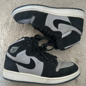 Nike Air Jordan 1 svart/grå sneakers - Snygga Nike Air Jordan 1 sneakers i svart och grått med klassisk swoosh-logga på sidan och Jumpman på hälen. Skorna har snörning, rund tå och platt sula. Ovandelen är i textil och mocka för en cool kontrast. Perfekta för dig som gillar streetstyle och ikoniska sneakers. Använda men fina.