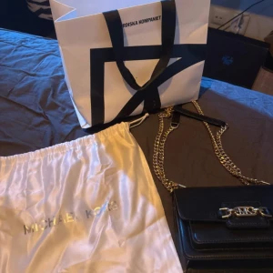 Svart Michael Kors axelväska i skinn - Snygg svart axelväska från Michael Kors i slätt skinn med guldfärgade kedjor och MK-detalj framtill. Väskan har ett stilrent, rektangulärt format och flera fack. Perfekt för dig som vill ha en trendig och praktisk väska med exklusiv känsla. Kvitto finns😊