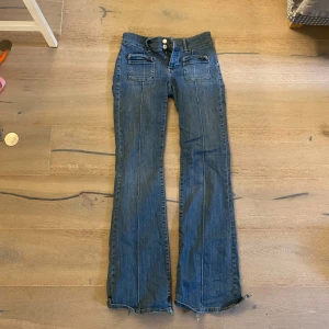 Blå bootcut jeans med fickdetaljer - Snygga blå jeans med bootcut passform och låg midja. Framtill finns två fickor med lock och knappar, samt markerade sömmar längs benen. Jeansen är en aning söndertrampade i slutet av bakre byxbenen, därav det låga priset. Nypris: 699