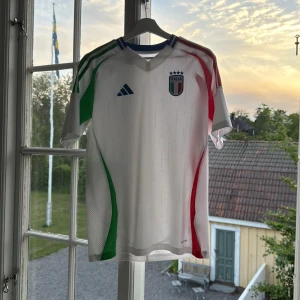 Italien adidas fotbollströja 15-16 år - Snygg vit Italien fotbollströja från adidas med gröna och röda detaljer på sidorna och axlarna. Tröjan har korta ärmar, blå adidas-logga och Italiens klubbmärke med fyra stjärnor på bröstet. Tillverkad i lätt och ventilerande material, perfekt för match eller träning.